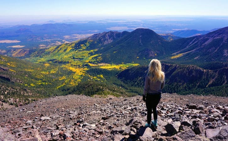 A woman stands atop a rocky overlook, looking down into a valley of green and yellow foliage surrounded by mountains