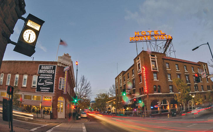 Dusk on a busy downtown street in a small town. Neon signs are on, one advertising the Hotel Monte Vista.