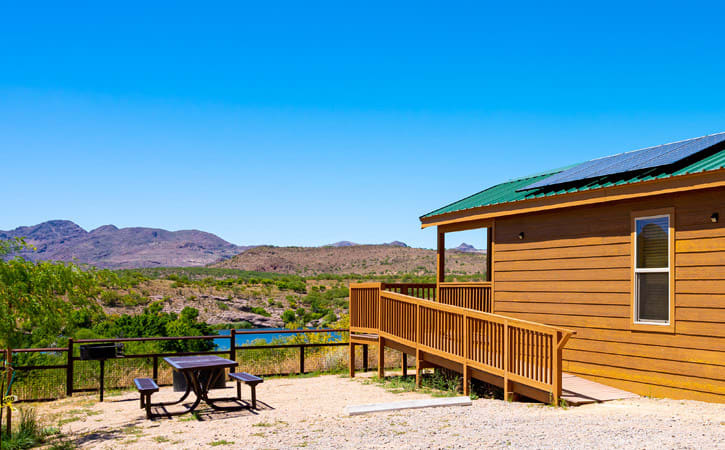 A wood cabin and metal picnic table overlook a landscape with a view of a lake, mountains and desert greenery