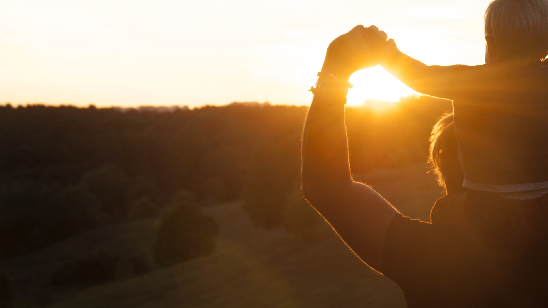 Silhouetted adult carrying a child on their shoulders at sunset, holding hands against a scenic backdrop of rolling hills and forest.